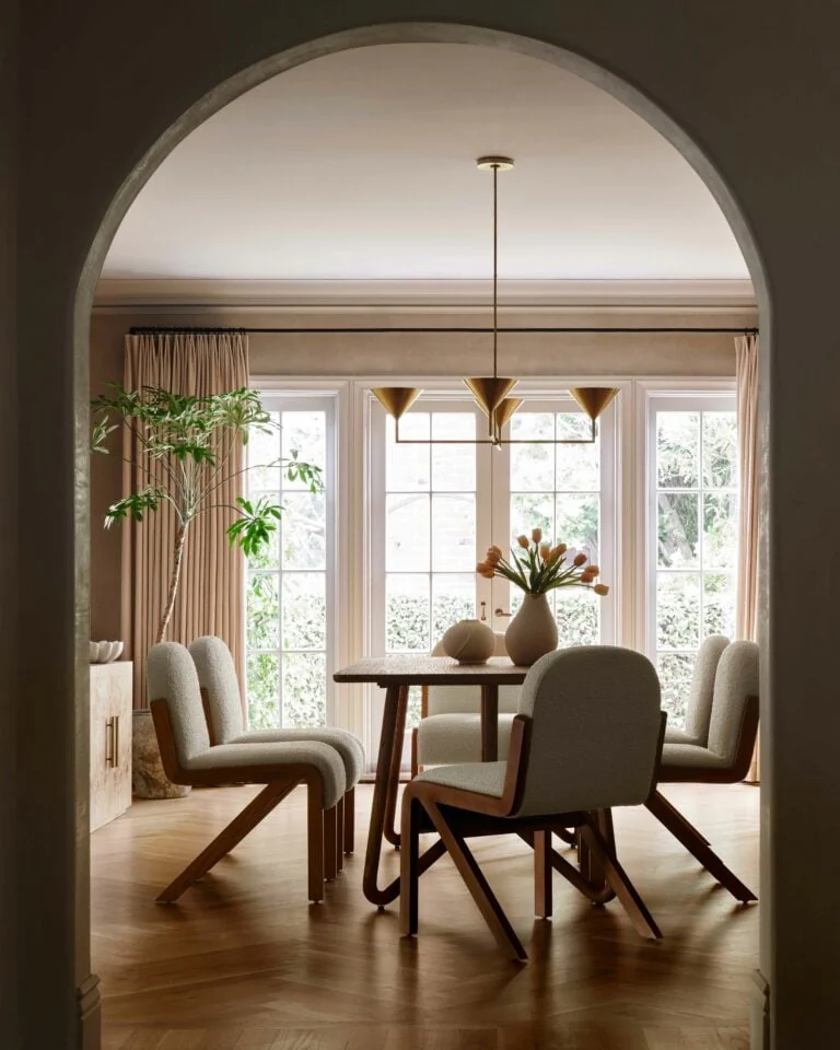 Dining room seen through an arch, bouclé chairs, wood table, brass pendant and tall windows on herringbone floor, for Anthropologie.