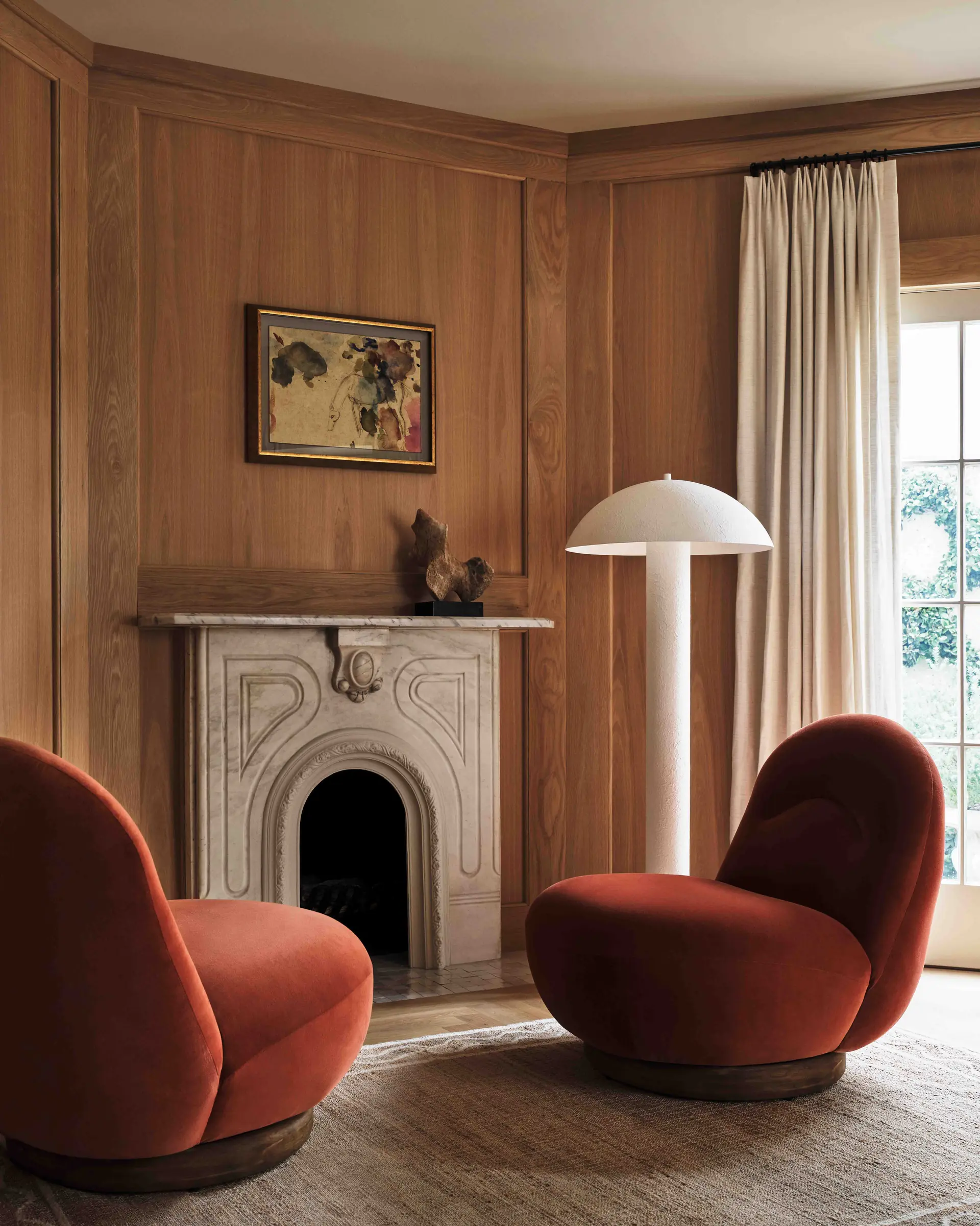 Two rust velvet swivel chairs by a carved stone fireplace in a wood-panelled room with a mushroom floor lamp, for Anthropologie.