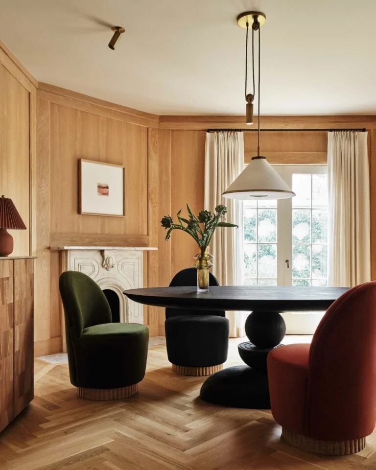 Dining room with oak paneling, black round pedestal table, green, black and rust velvet chairs and marble fireplace, for Anthropologie.