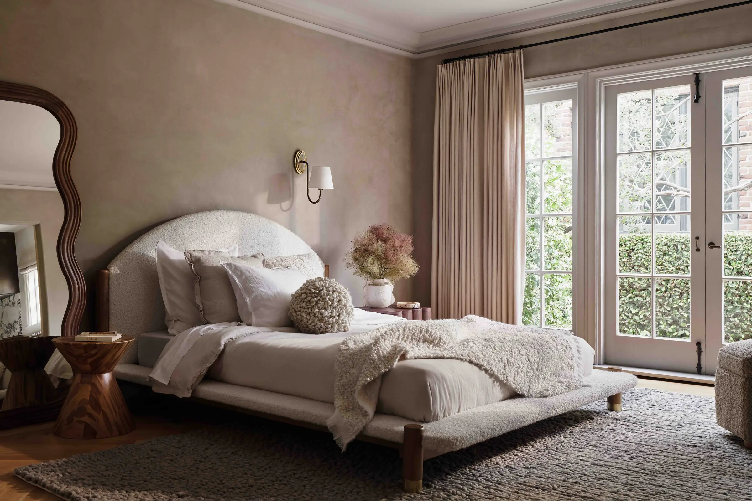 Bedroom interior with arched boucle bed, wavy wood mirror, linen drapes and French doors to a garden, for Anthropologie.