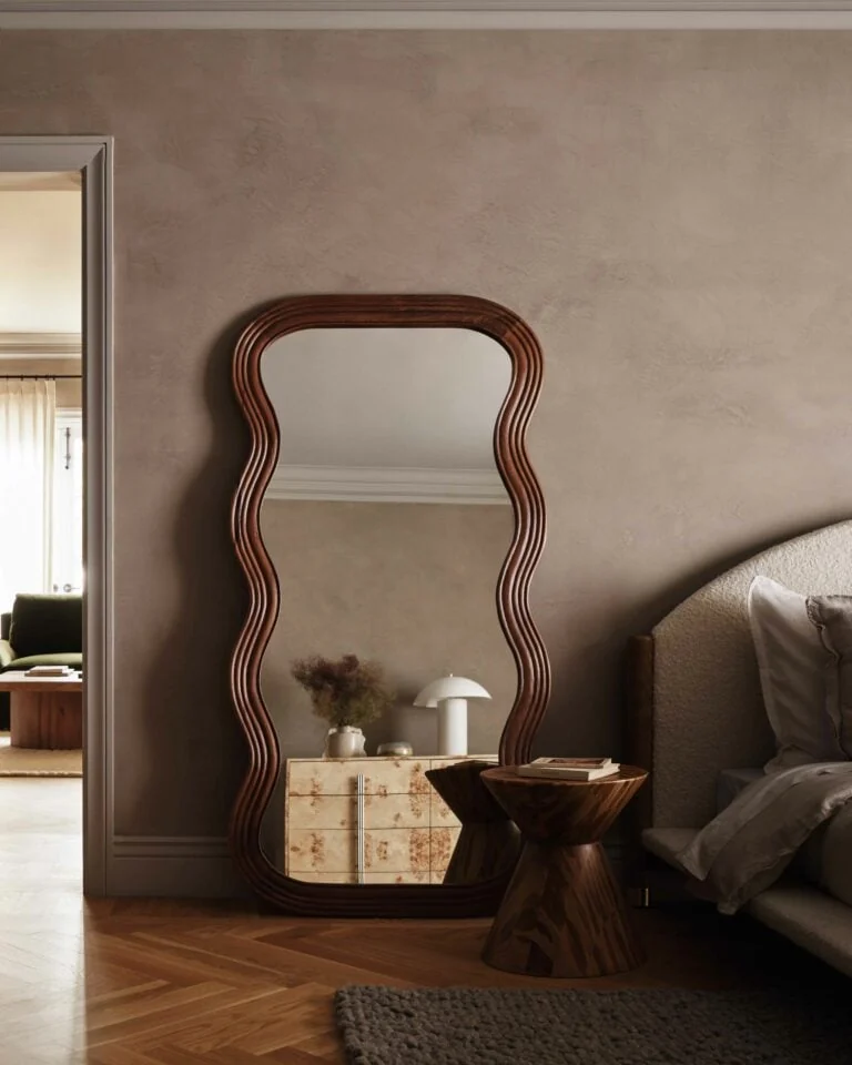Wavy walnut floor mirror leaning in a bedroom beside a carved wood side table and upholstered bed, for Anthropologie.