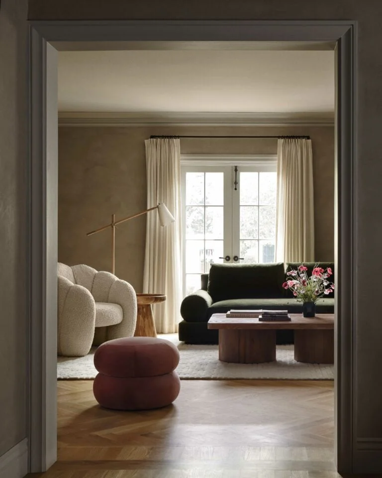 Living room seen through a doorway, with bouclé chair, green velvet sofa, wood coffee table and pink pouf, for Anthropologie.