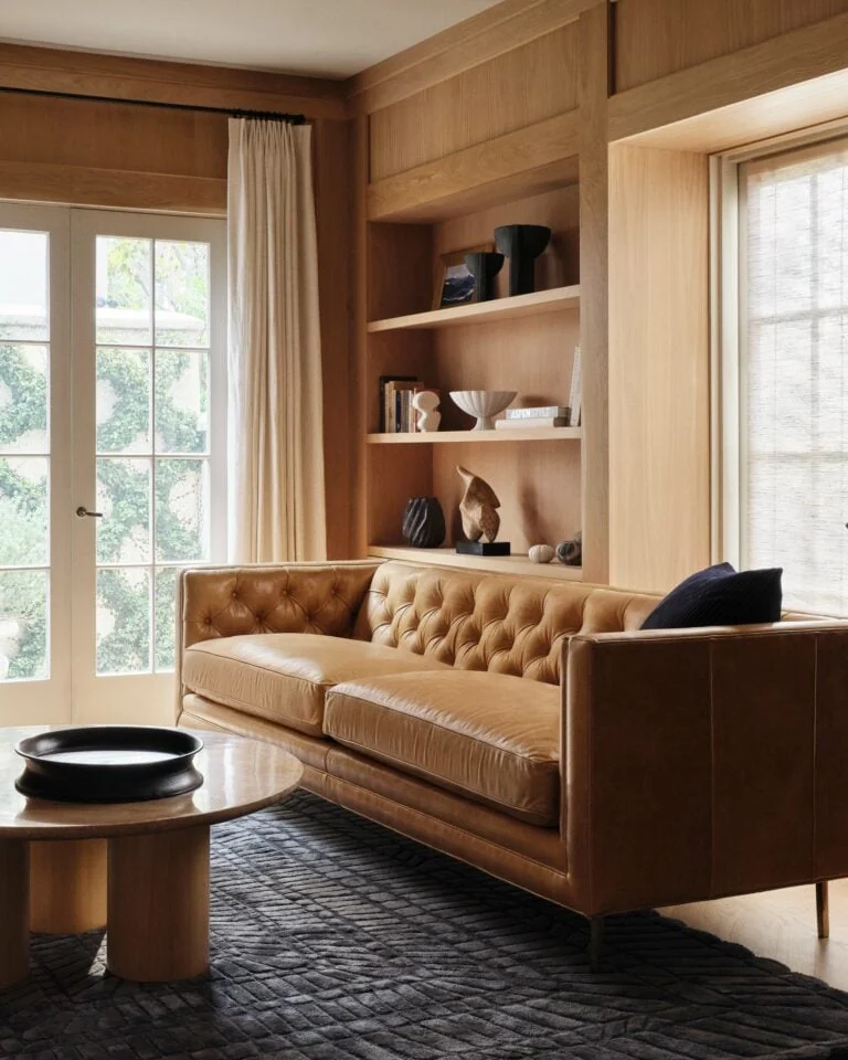 Tan tufted leather sofa in a wood-panelled living room with built-in shelves and a round coffee table, for Anthropologie.