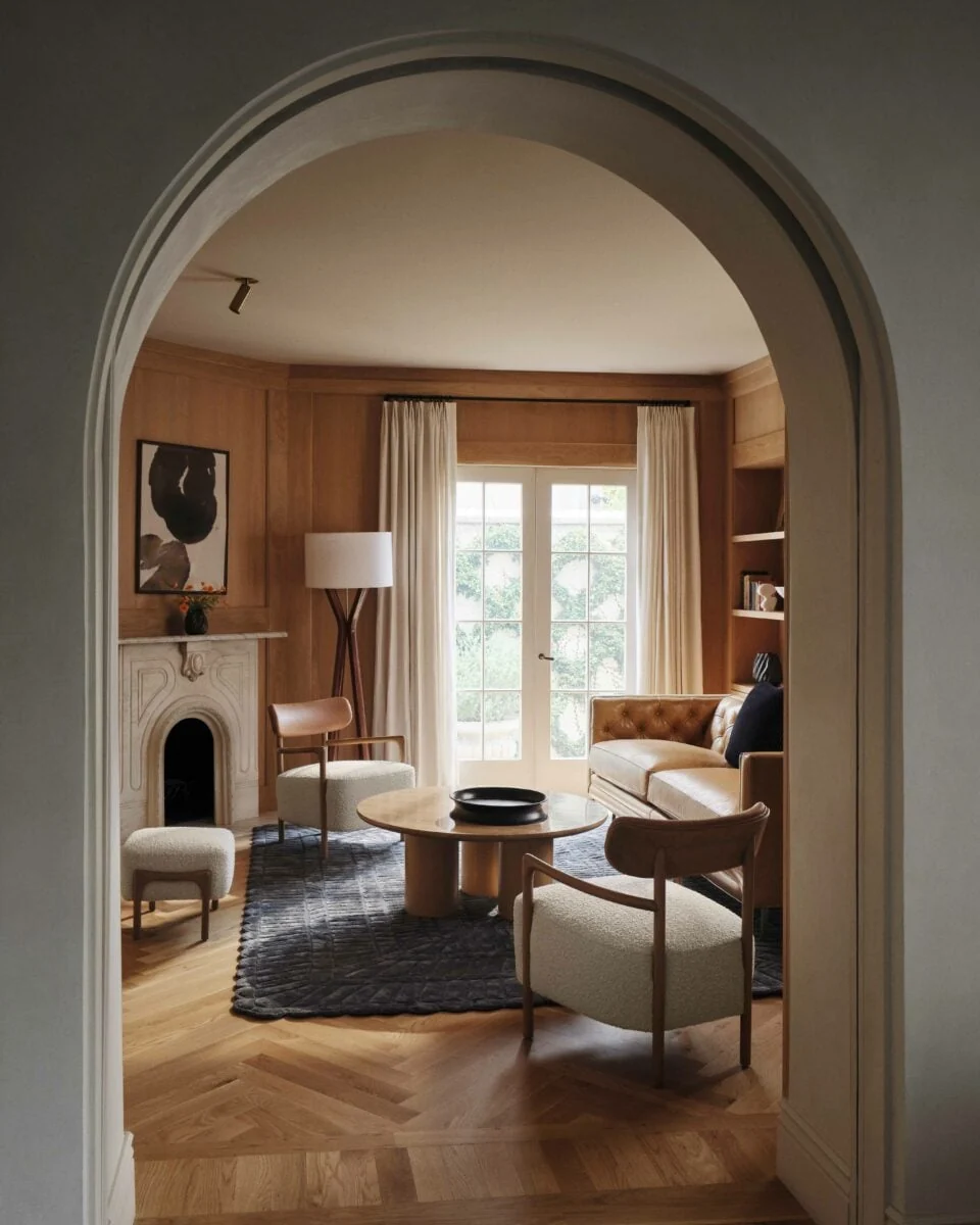 Living room seen through an arched doorway, with oak paneling, marble fireplace, boucle chairs and leather sofa, for Anthropologie.