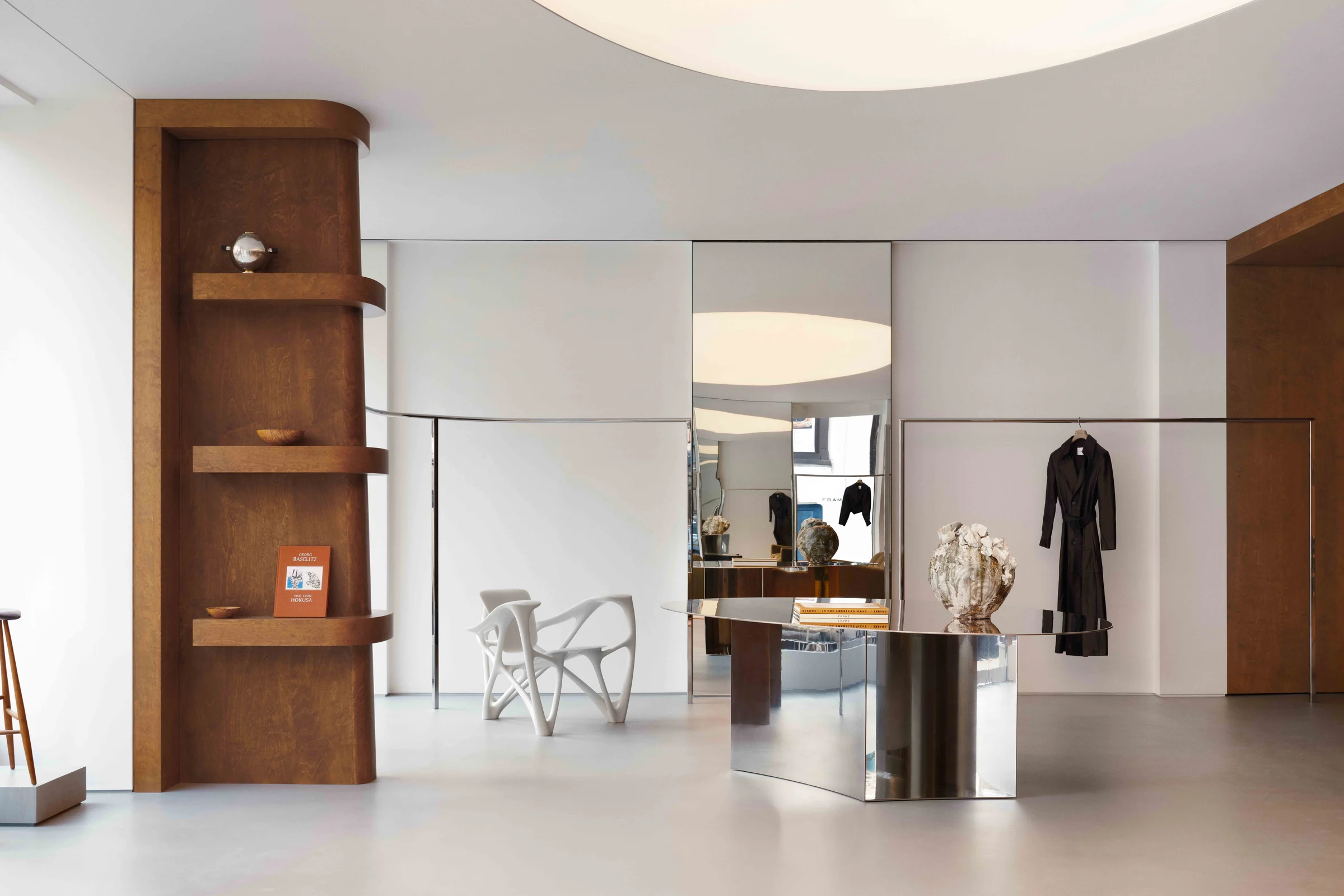 Boutique interior with oak shelving, sculptural white chair, mirrored steel counter and black coat on a rail, for Frame.