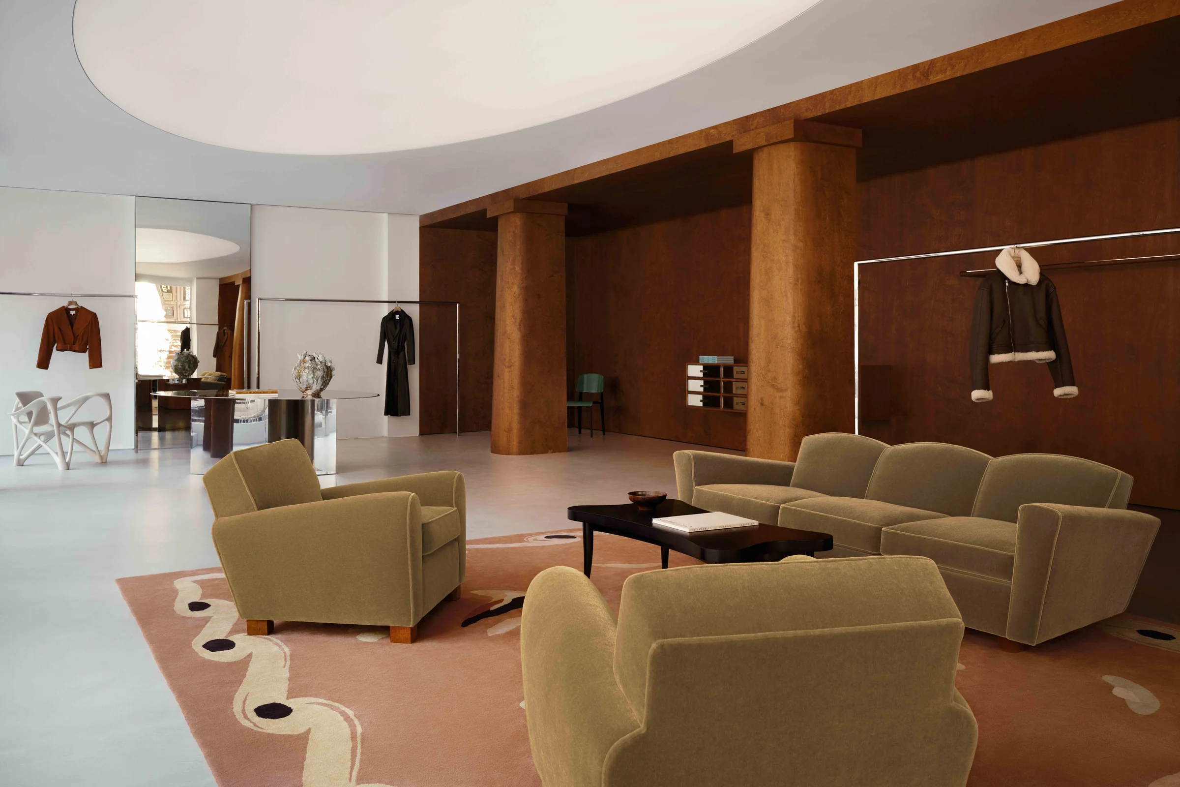 Boutique interior with olive velvet sofa and armchairs, wood-panelled columns and racks of coats, for Frame.