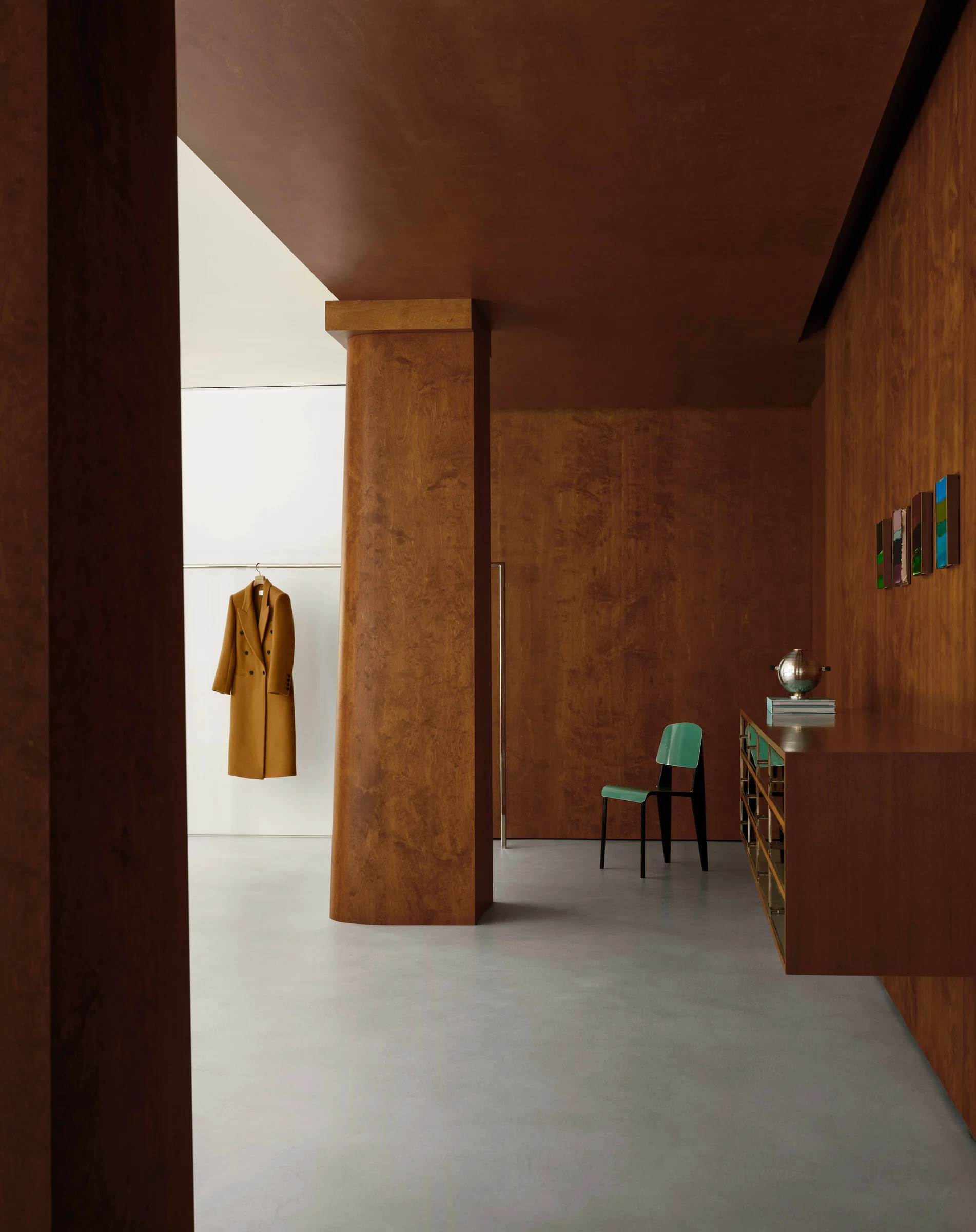 Boutique interior with wood-paneled walls, a camel coat on a rail and a green chair on grey concrete, for Frame.