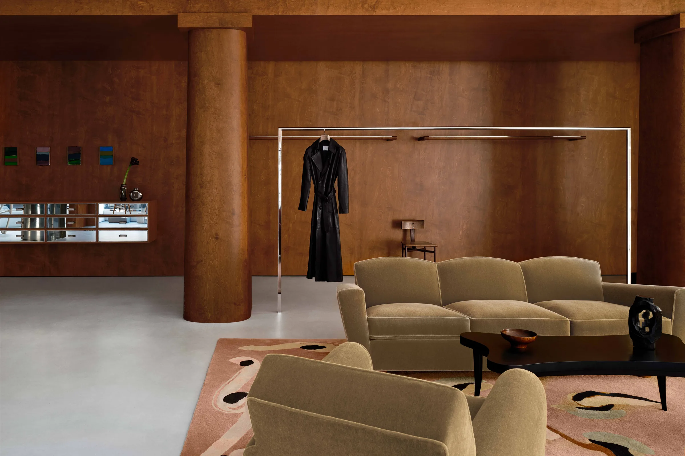Boutique interior with wood-panelled walls, velvet sofa, black leather trench on a chrome rail and abstract rug, for Frame.