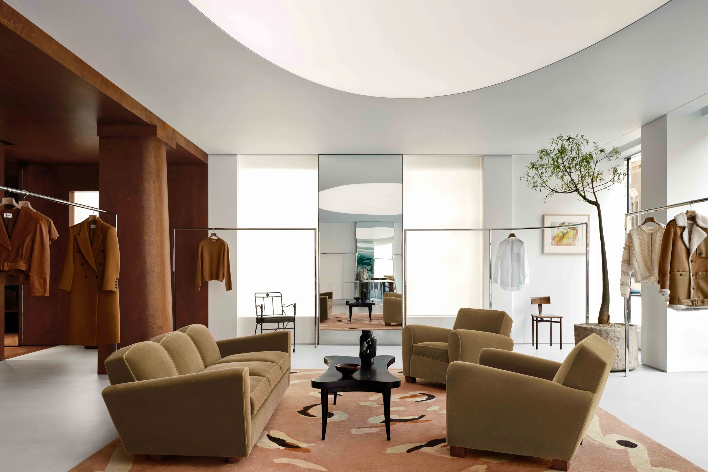 Boutique interior with olive velvet sofa and armchairs, patterned rug and coats on chrome rails under a curved ceiling, for Frame.