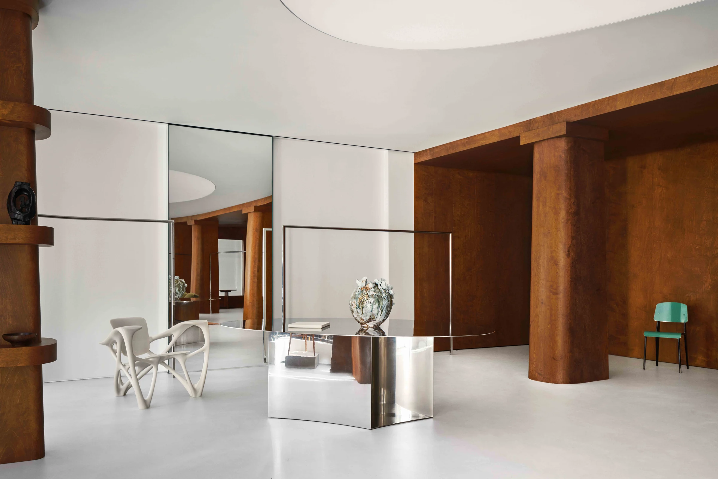 Interior with burl wood columns, a mirrored steel desk, sculptural white chair and green chair on a white floor, for Frame.