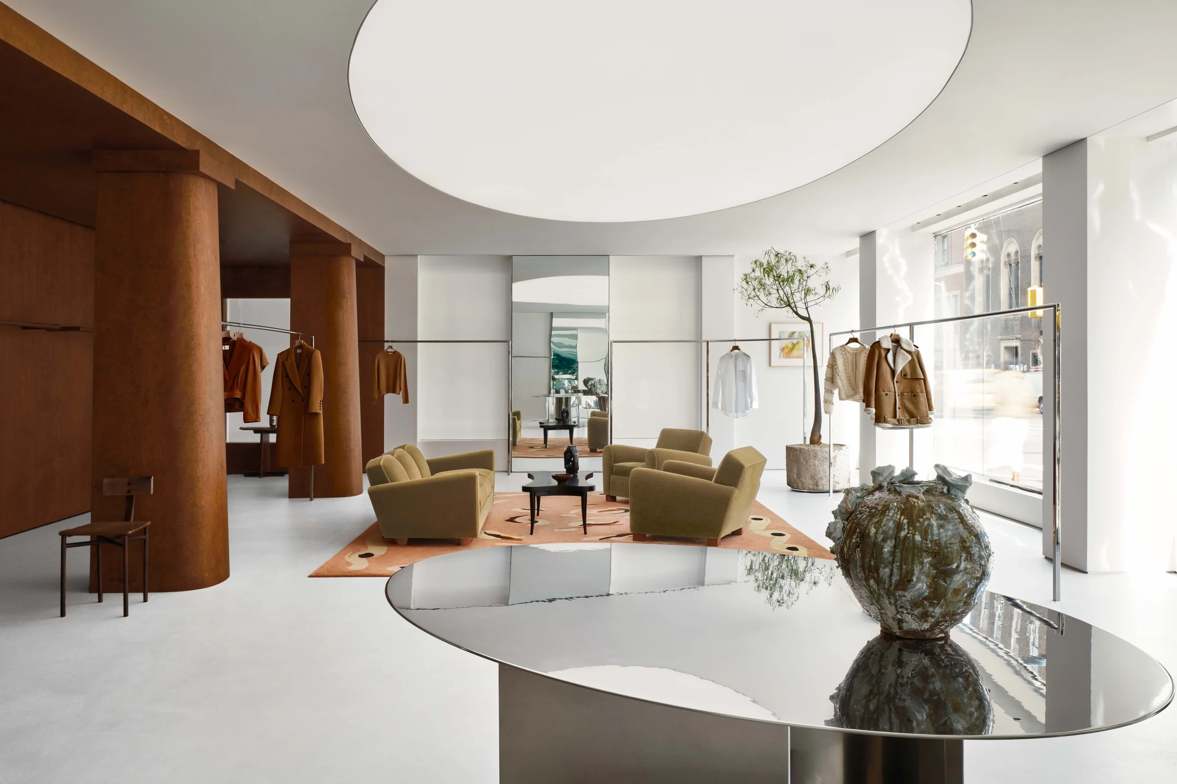 Retail store interior with olive velvet armchairs, rust columns, coats on rails and a mirrored table, for Frame.