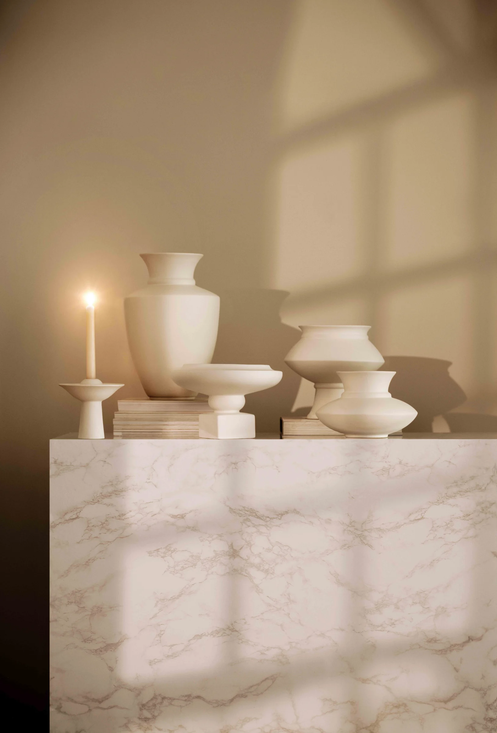 White ceramic vases and a lit candle on a marble plinth with window shadows on a beige wall, for H&M Home.