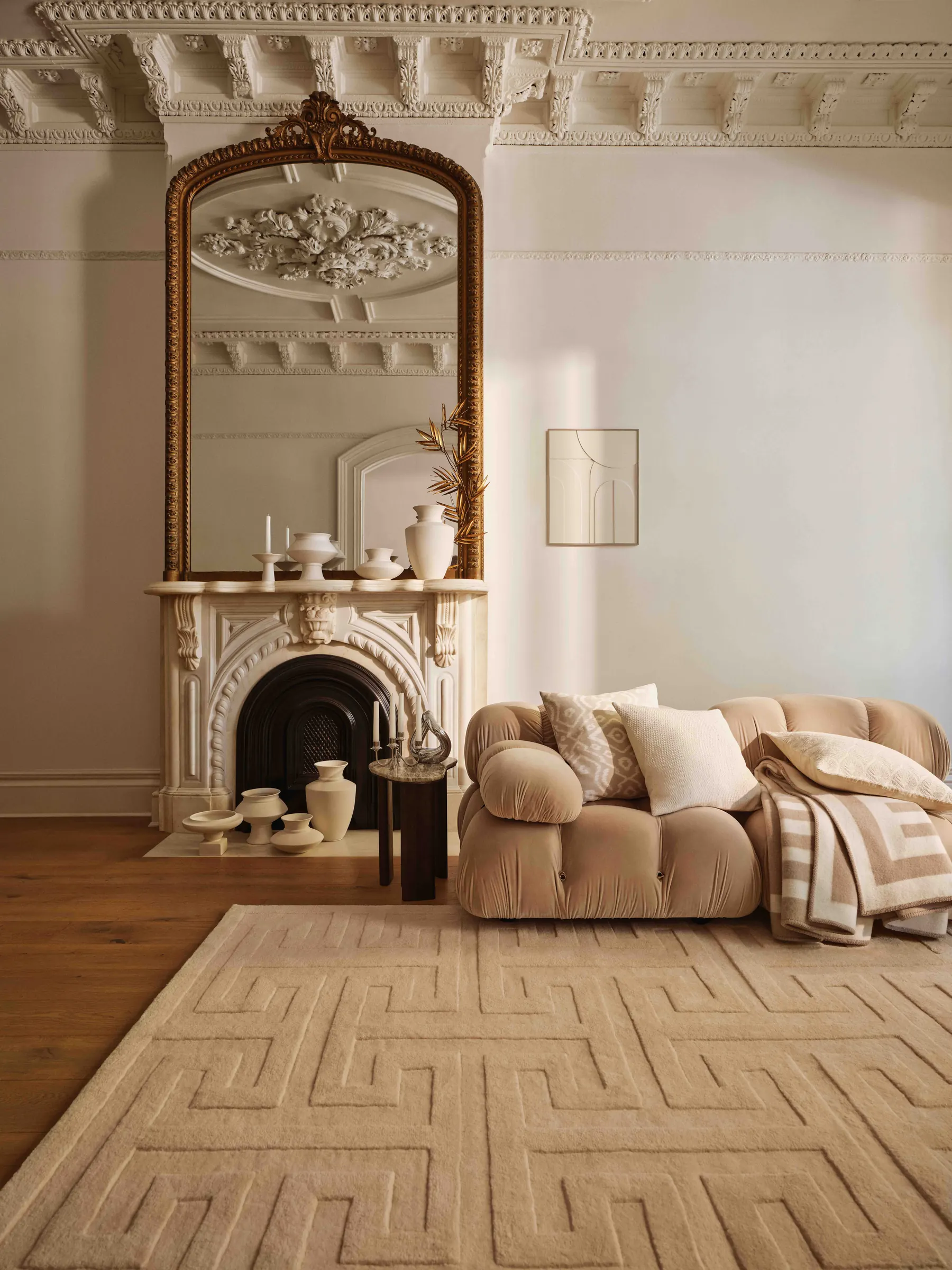 Living room with beige velvet sofa, carved marble fireplace, gilded mirror and geometric wool rug, for H&M Home.