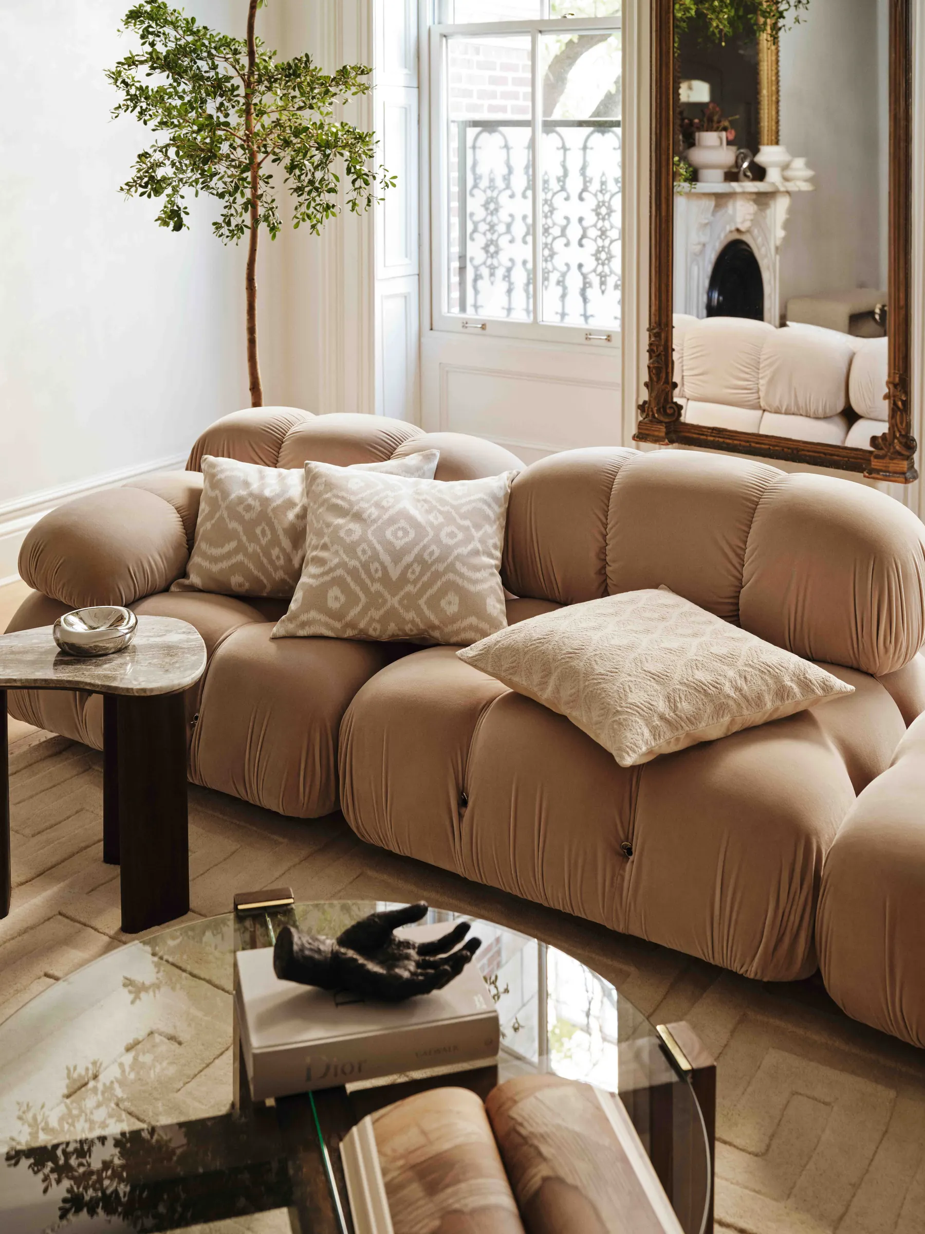 Living room with a tan velvet modular sofa, patterned cushions, glass coffee table and gilded mirror, for H&M Home.
