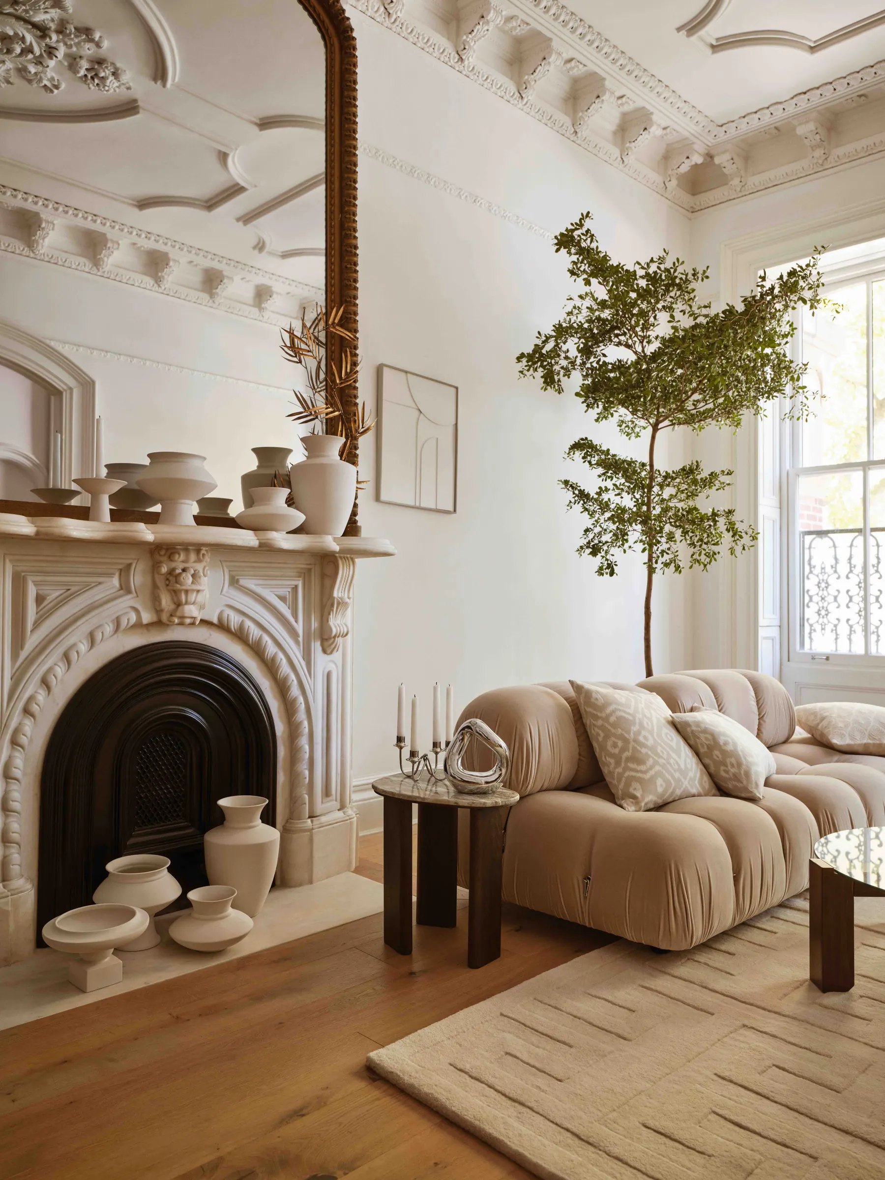 Ornate white parlor with marble fireplace, ceramic vases, beige modular sofa and olive tree by the window, for H&M Home.