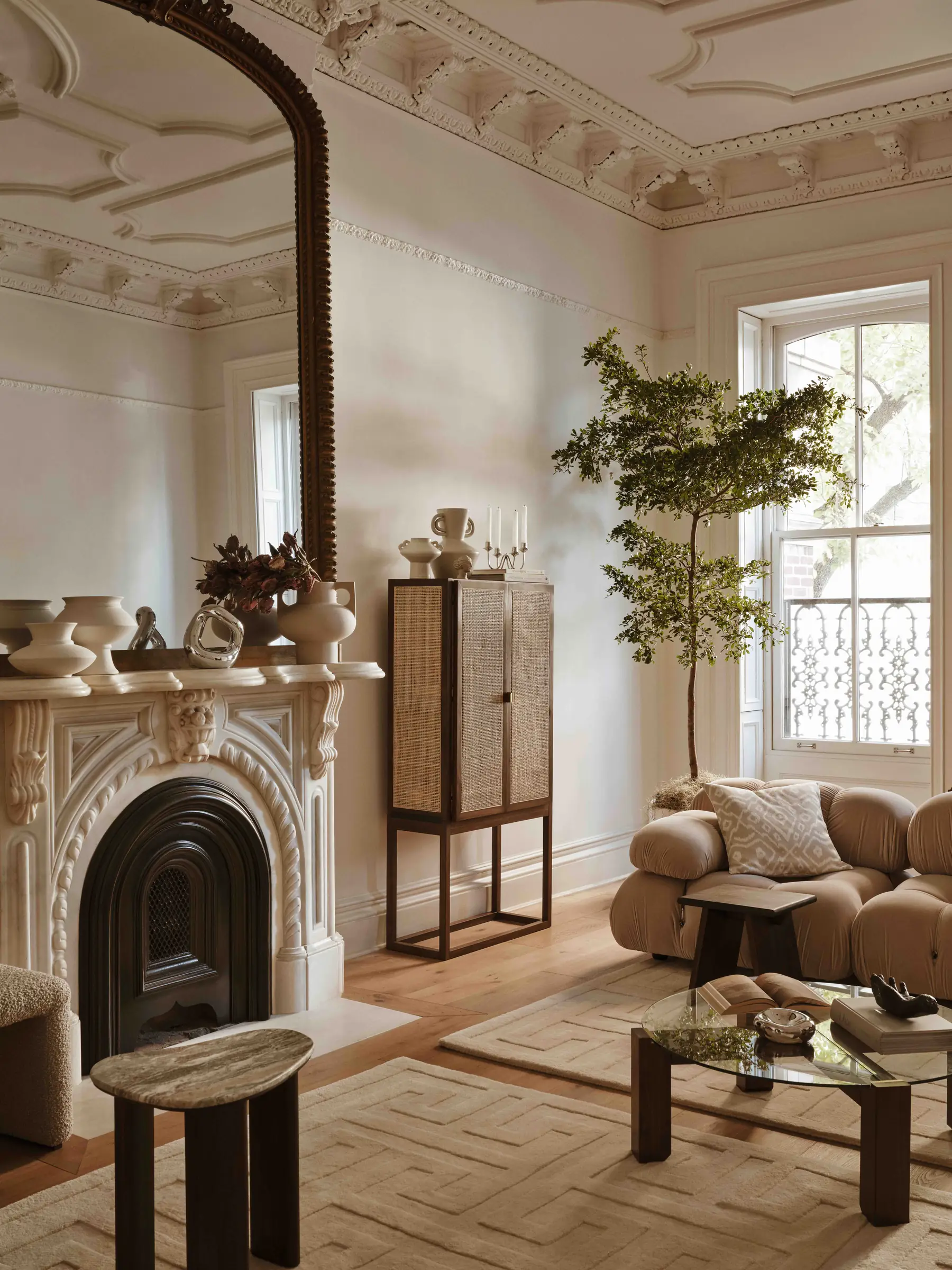 Period living room with carved white fireplace, rattan cabinet, olive tree and curved bouclé sofa, for H&M Home.