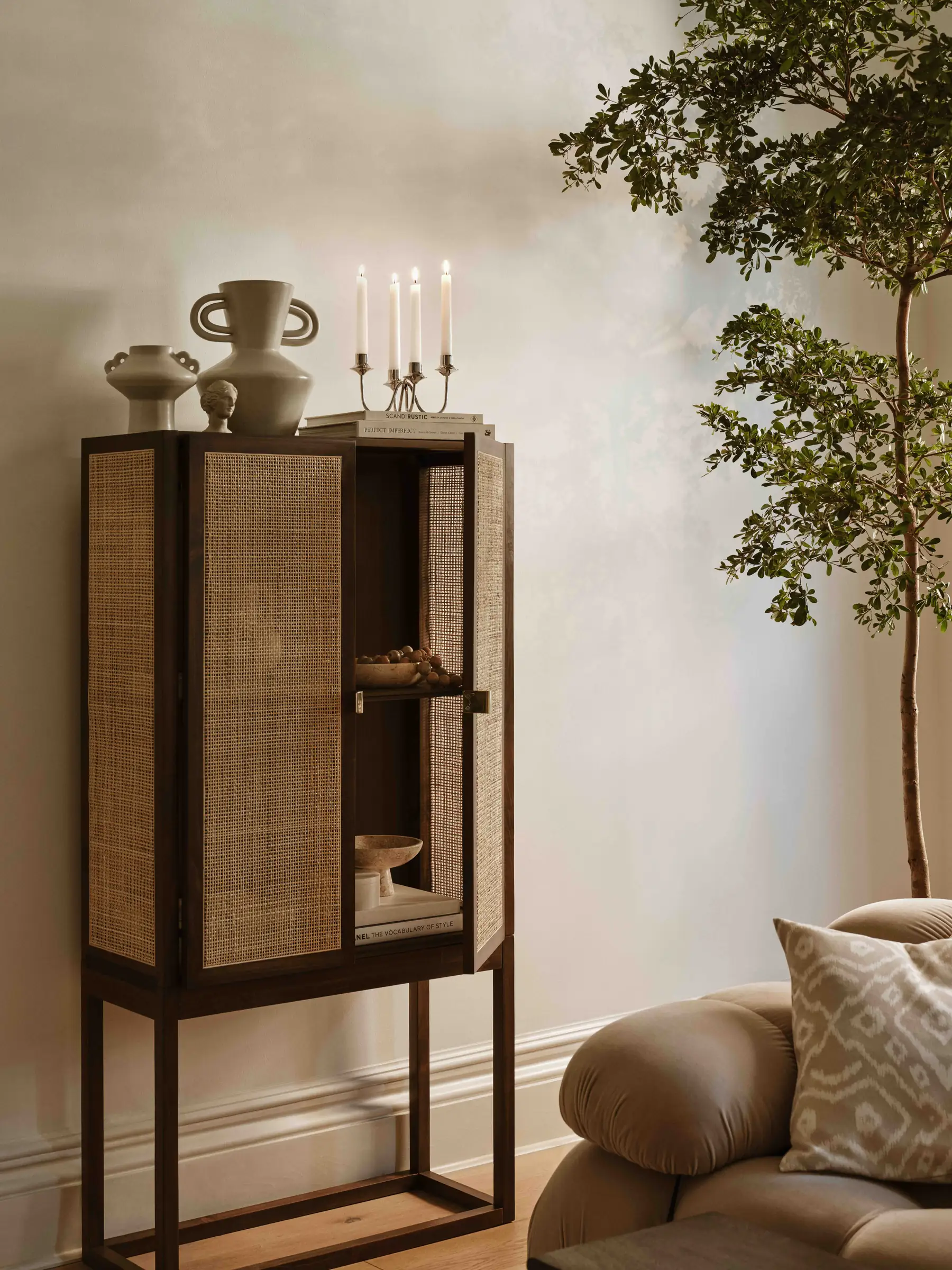 Dark wood rattan cabinet with vases and lit candelabra beside an olive tree and curved sofa, for H&M Home.