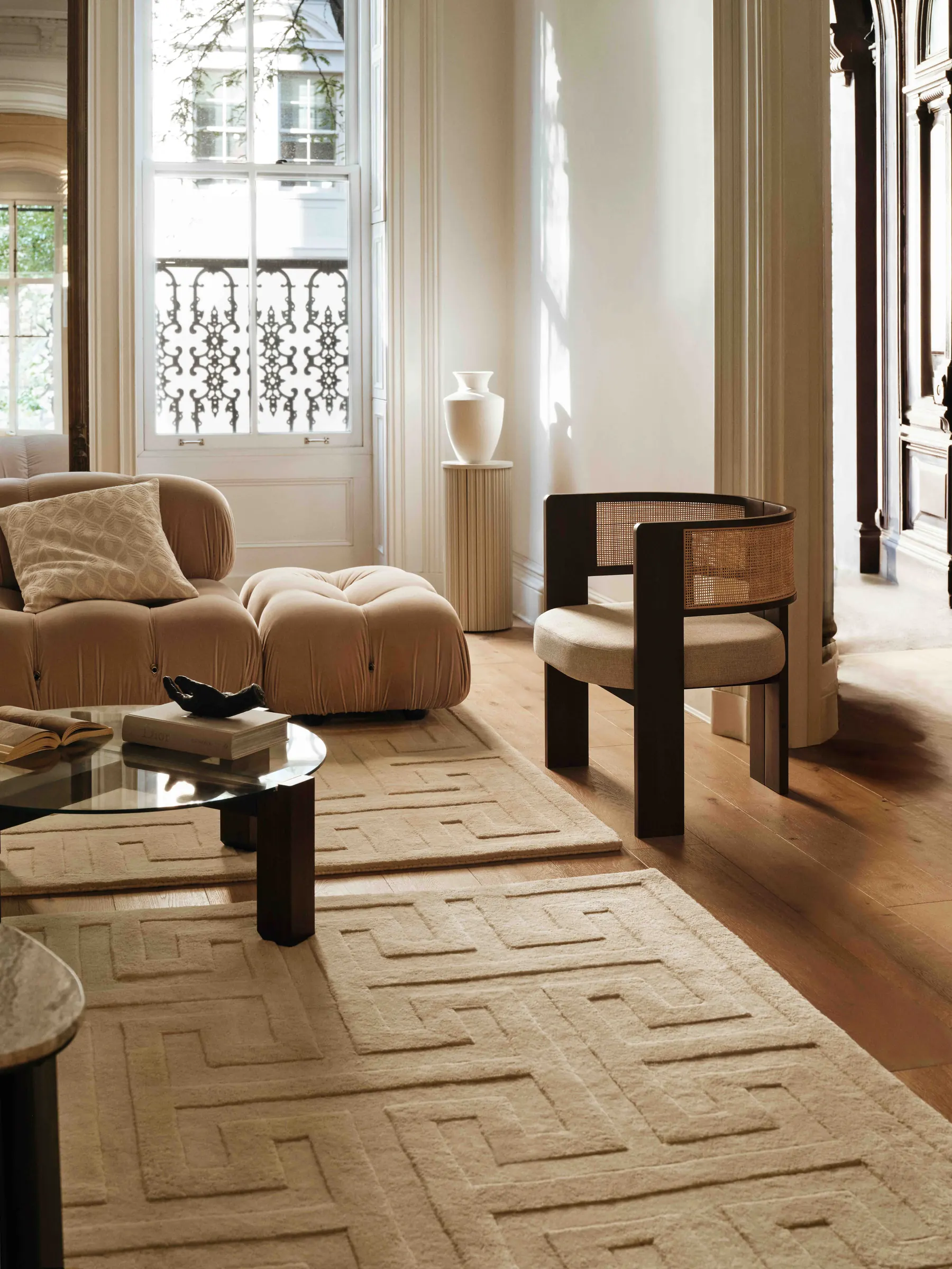 Living room interior with cream key-pattern rug, velvet sofa, cane armchair and glass coffee table, for H&M Home.
