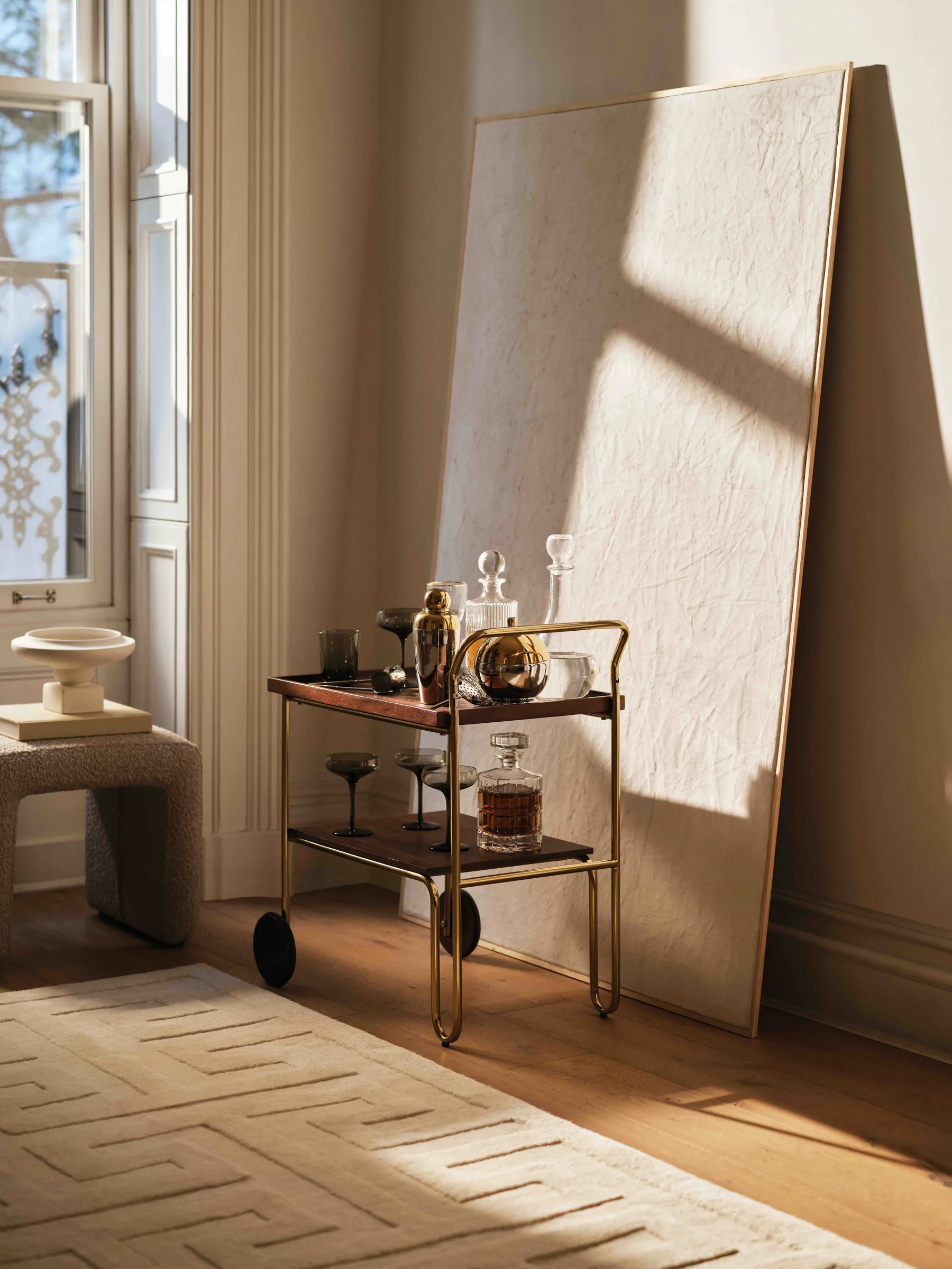 Brass bar cart with decanters and coupe glasses beside a leaning textured canvas in a sunlit room, for H&M Home.