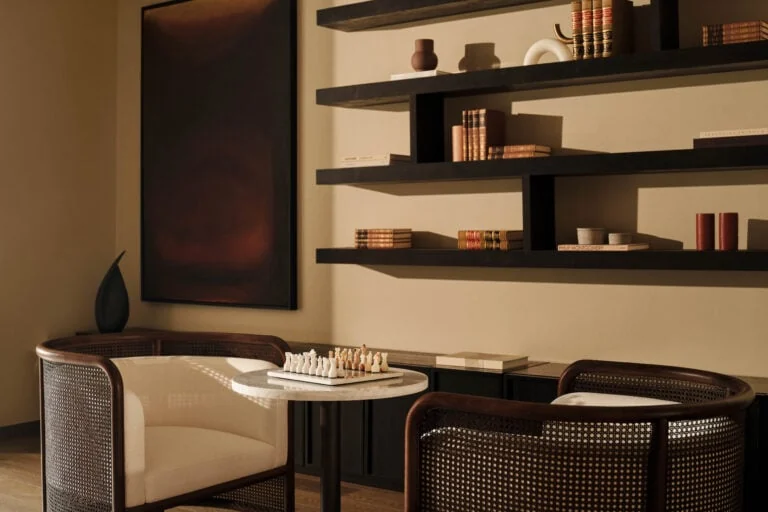 Lounge corner with cane armchairs, marble chess table and dark shelves of books under warm light, for Aman.