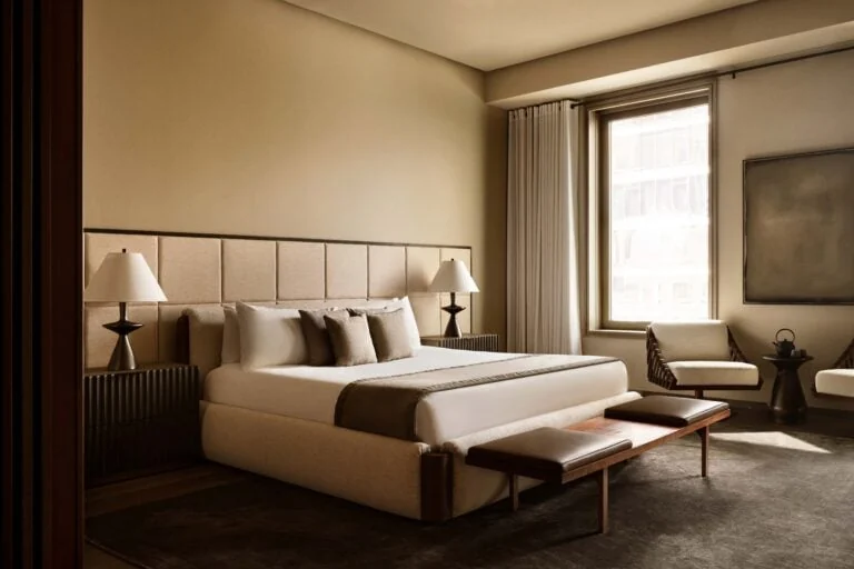 Hotel suite with a cream bed, paneled headboard, leather bench and armchairs by a tall window, for Aman.