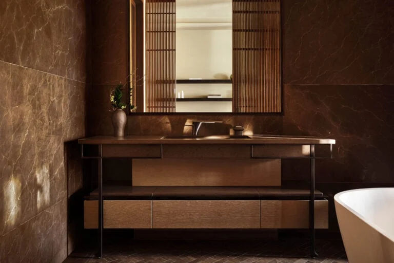Hotel bathroom with brown marble walls, wood and metal vanity, framed mirror and freestanding tub, for Aman.