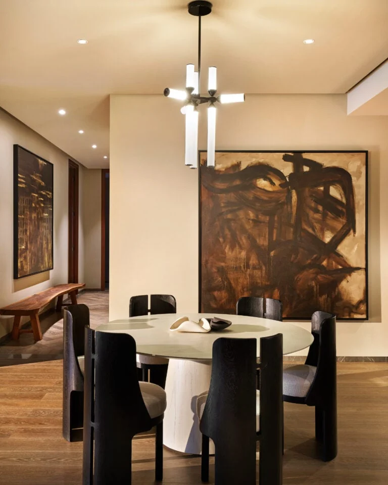 Dining room with round stone table, dark wood chairs, tube chandelier and large brown abstract painting, for Aman.