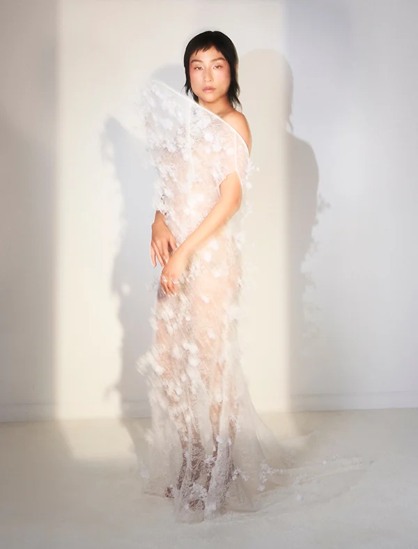 Model in a sheer white one-shoulder lace gown with 3D floral appliqués, standing in sunlight against a white wall, for Loewe.