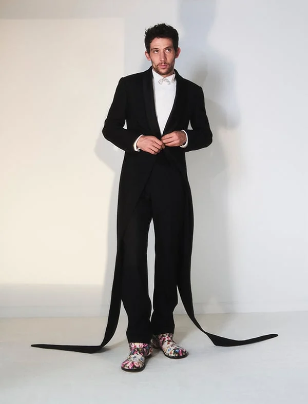 Man in a black tailcoat with long trailing ribbon tails and floral print shoes in a white studio, for Loewe.
