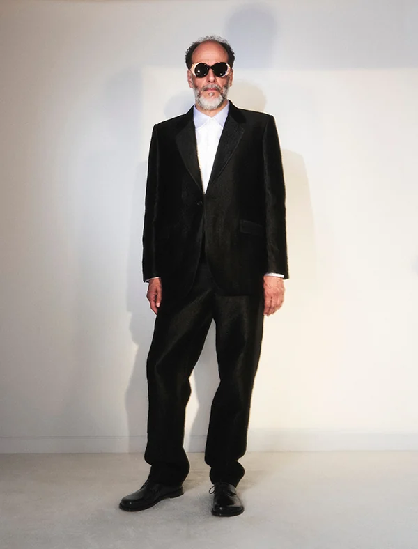 Bearded man in a black satin suit, white shirt and round dark sunglasses standing against a white wall, for Loewe.