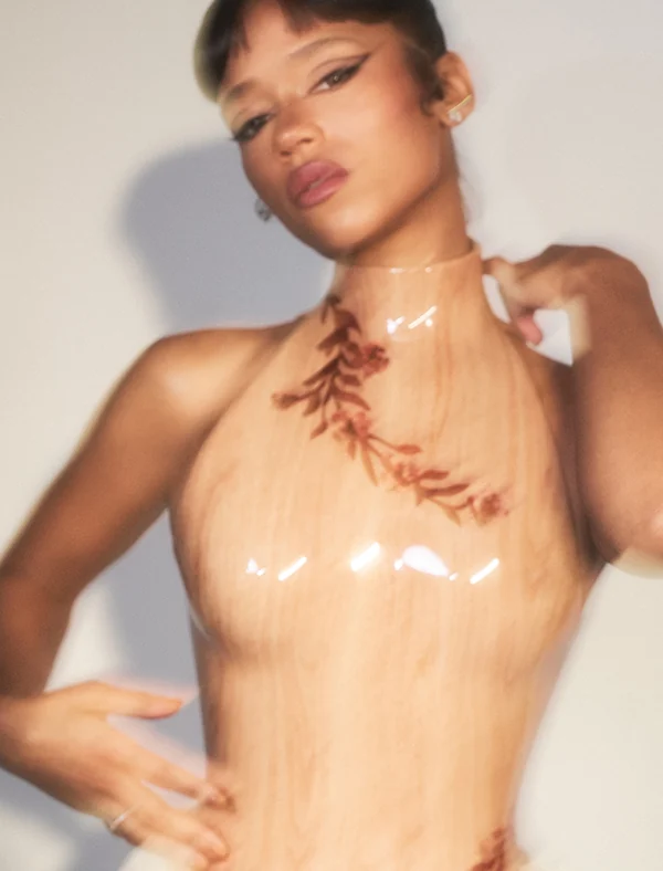 Model in a glossy wood-grain halter top with leaf print, posing against a pale wall with soft motion blur, for Loewe.
