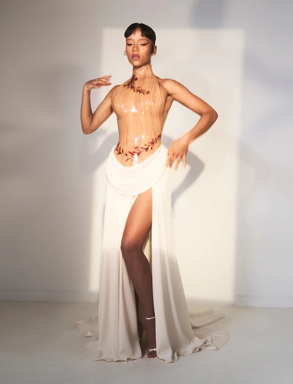Model in an ivory halter gown with a sheer sculpted bodice, leaf motifs and high slit, posed in soft sunlight, for Loewe.