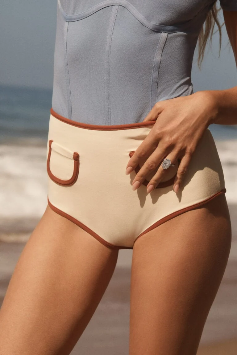 Model on a beach wearing an oval diamond ring, hand resting on cream high-waist swim shorts, for Norman Silverman.